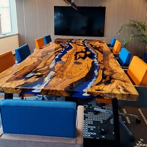 Puede incluir: Una gran mesa de conferencias hecha a medida con un diseño único. La mesa presenta una superficie de madera con una incrustación de resina epoxi azul en forma de río. La mesa está rodeada de sillas azules y naranjas. La mesa mide aproximadamente 305 cm de largo.