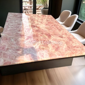 May include: A rectangular table with a polished, pinkish-red surface, possibly stone. The table is supported by a dark base and surrounded by white chairs. The surface has a unique, natural pattern. The text "ADAMSTONEShop" is visible on the table.