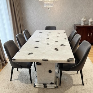 May include: A white marble dining table with dark grey inclusions, supported by a matching pedestal base. Eight grey upholstered chairs with black legs surround the table. A crystal chandelier hangs above, illuminating the dining space. Table dimensions in cm.