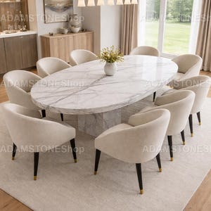 May include: An oval marble dining table with a matching marble base is surrounded by ten cream-colored upholstered chairs. The chairs have black legs with gold accents. A vase of flowers sits on the table, and the room has a neutral color palette.