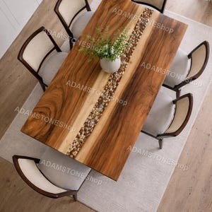 Op de afbeelding: Vogelperspectief van een houten eettafel met een centrale rivier van stenen en een vaas met groen. De tafel is omringd door zes stoelen met lichtgekleurde kussens en geweven rugleuningen. De tafel staat op een lichtgekleurd tapijt.