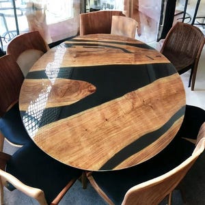 Può includere: Un tavolo da pranzo rotondo in legno con un inserto in resina epossidica nera. Il tavolo ha una finitura in legno naturale ed è circondato da sei sedie con rivestimento nero.