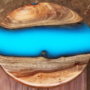 May include: Round wooden table top with a blue epoxy resin inlay. The wood has a natural brown colour and a wood grain pattern.