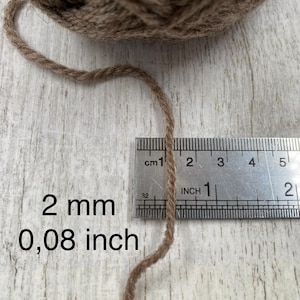 May include: A ball of brown yarn with a single strand extending downward. A metal ruler is positioned next to the yarn, showing a thickness of 2 mm or 0.08 inches. The text 2 mm and 0.08 inch is printed on the left side of the image.