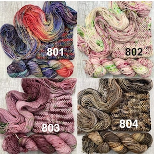 May include: Four sets of hand-dyed yarn skeins and matching knitted swatches labelled 801, 802, 803, and 804. Set 801 features vibrant red, purple, and blue tones. Set 802 is cream with green and pink speckles. Set 803 is dusty pink and mauve. Set 804 is brown and tan.