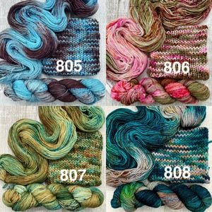 May include: Four sets of hand-dyed yarn skeins and matching knitted swatches labelled 805, 806, 807, and 808. The yarn colours include blue and brown, pink and olive, green and gold, and teal and dark blue, each featuring variegated colour patterns.