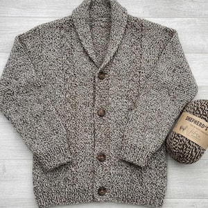 May include: A brown and white speckled knit cardigan featuring a shawl collar, long sleeves, and four dark brown buttons down the front. The garment is crafted with a cable knit pattern along the front panels and includes a ball of matching yarn.