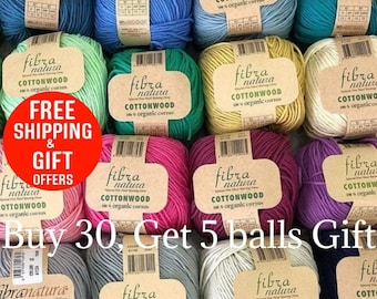 May include: A collection of Fibra Natura Cottonwood yarn balls in various colours, including blue, green, yellow, pink, purple, grey, and white. Each ball features a tan paper label with brand text and organic cotton specifications.