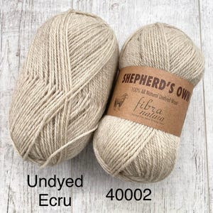 May include: Two skeins of light beige, undyed wool yarn. One skein features a brown paper label with the text Shepherd's Own, 100% All Natural Undyed Wool, Fibra Natura, and the image of a sheep. The text Undyed Ecru and 40002 is visible.