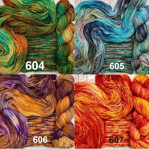 May include: Four skeins of hand-dyed yarn with matching knitted swatches labelled 604, 605, 606, and 607. The yarn colours include green and orange, blue and purple, purple and gold, and vibrant red and orange variegated tones.