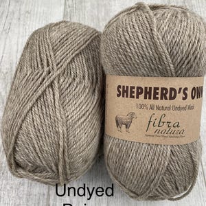 May include: Two skeins of beige undyed wool yarn. The right skein features a tan paper label with the text SHEPHERD'S OWN, 100% All Natural Undyed Wool, and fibra natura, accompanied by a small illustration of a sheep. The text Undyed Beige 40003 is visible.