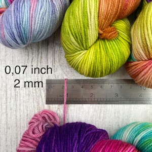 May include: Multiple skeins of multi-coloured yarn in shades of pink, purple, blue, orange, and lime green. A metal ruler is positioned against a single strand of pink yarn, showing a thickness of 0.07 inches and 2 mm.