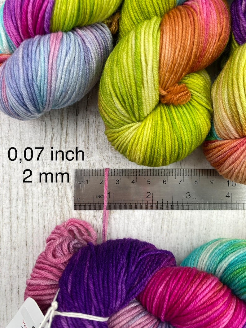 May include: Multiple skeins of multi-coloured yarn in shades of pink, purple, blue, orange, and lime green. A metal ruler is positioned against a single strand of pink yarn, showing a thickness of 0.07 inches and 2 mm.