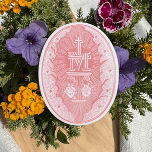 May include: Oval sticker with a pink background and white graphic. The graphic features a cross, the letter M, two hearts, and other religious symbols. The sticker is surrounded by greenery and colourful flowers.