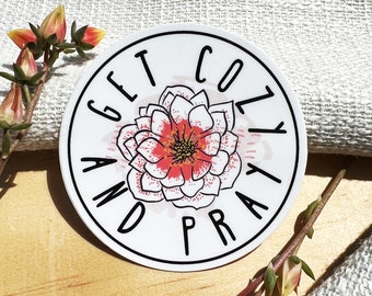Puede incluir: Una pegatina redonda blanca con borde negro. El texto "GET COZY AND PRAY" está impreso en negro. Un diseño floral con pétalos rojos y blancos está en el centro. La pegatina está sobre una superficie de madera con detalles florales.