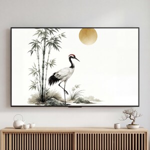 Puede incluir: Una impresión artística enmarcada que representa una grulla y bambú, con un sol dorado. La grulla tiene una corona roja, plumas negras y blancas y un pico largo. La obra de arte se exhibe en una pared sobre un mueble de madera.
