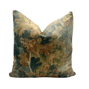 Puede incluir: Un cojín cuadrado de tela texturizada con un denso patrón floral multicolor en tonos azul verdoso apagado, verde bosque profundo y marrón dorado cálido. El cojín tiene un aspecto suave y mullido con un pliegue central visible.