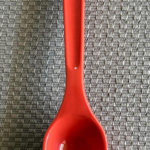 May include: A red plastic spoon with a unique handle design. The spoon is on a gray textured surface.