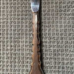 May include: A silver butter knife with a decorative handle. The handle has a scalloped design and a rounded end.