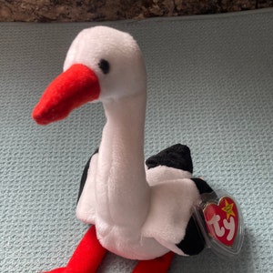 May include: White and black stuffed stork with red beak and legs. The stork is sitting on a light blue surface. The Ty Beanie Baby tag is attached to the stork's back.