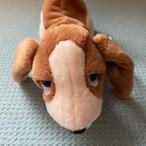 May include: A brown and white plush toy dog with floppy ears and a black nose. The dog is lying on its belly with its head tilted down.