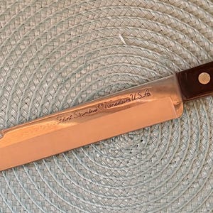 May include: A long, stainless steel carving knife with a brown wooden handle. The blade is a light, reflective color, and the handle is secured with two rivets. The knife has text on the blade that reads "Karl Schmidt, Solingen USA."