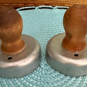 May include: Two vintage metal biscuit cutters with wooden handles. The cutters have a circular shape with a small hole in the center. The wooden handles are a light brown color. The cutters are sitting on a light blue surface.