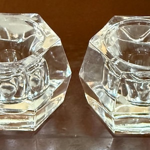 May include: Two clear, faceted glass candle holders. Each holder has a hexagonal shape with a central well for a candle. The glass is transparent, allowing light to pass through. The candle holders are displayed on a dark surface.