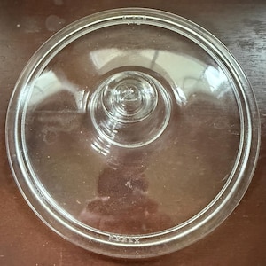 May include: A clear, circular glass lid featuring a central knob handle. The rim is slightly raised, and the glass is embossed with the text PYREX and the number 623-C near the edge. The design is simple, functional, and transparent.