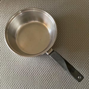 May include: Stainless steel saucepan with a black handle. The pan has a round shape and a silver finish. The handle is black and has a twisted design. The pan is sitting on a gray textured surface.