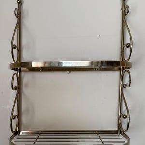 May include: A two-tiered, vintage-style wall shelf with a metallic finish. The shelf has a curved design with decorative scrollwork and a slatted bottom shelf. The top shelf is solid. The overall design is ornate and elegant.