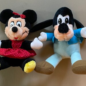 May include: A plush Mickey Mouse doll wearing a red dress with white polka dots and a yellow bow.  A plush Goofy doll wearing a blue shirt and pants.