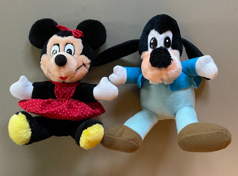 May include: A plush Mickey Mouse doll wearing a red dress with white polka dots and a yellow bow.  A plush Goofy doll wearing a blue shirt and pants.