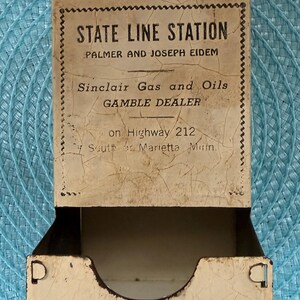 May include: Vintage match holder with the text "STATE LINE STATION" and "Sinclair Gas and Oils." The cream-colored metal container shows signs of age, with a hanging loop at the top and a curved opening at the bottom.