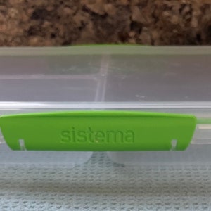 May include: A clear plastic Sistema lunch box with a green clip closure. The rectangular container has three compartments inside. The word "sistema" is printed in white on the green clip.