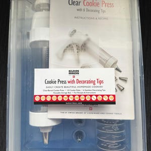 May include: A clear plastic cookie press kit with a white plunger, decorating tips, and cookie disks. The packaging includes instructions and recipes. The product is designed for making homemade cookies.