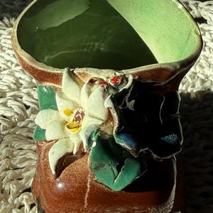 May include: A brown ceramic boot-shaped planter with a glossy finish. The front features a three-dimensional white flower with yellow centers and green leaves. The interior of the boot is glazed in a light green color.