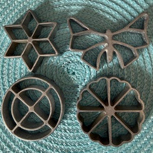 May include: Four vintage metal cookie cutters in various shapes: a six-pointed star, a butterfly, a circle with crossbars, and a scalloped flower. The cutters are made of a silver-colored metal and are arranged on a turquoise woven mat.