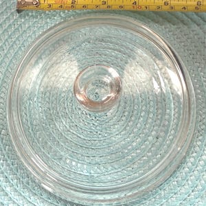 May include: A clear glass lid with a circular design and a small, rounded handle. The lid is approximately 6 inches in diameter. The lid is on a light blue woven surface. A yellow measuring tape is in the background.