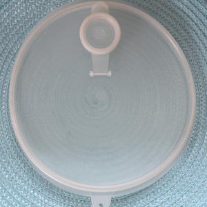 May include: A clear, round plastic lid with a handle and a small tab. The lid is designed to fit a container and has a circular shape. The handle is centered and the tab is on the side.