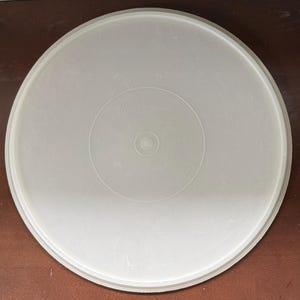 May include: A round, off-white Tupperware lid. The lid has a slightly textured surface and a raised edge. The word "Tupperware" is embossed in the center. The lid is designed to fit a container.
