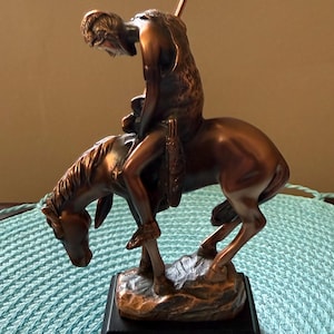 May include: Bronze-colored statue depicting a Native American on horseback, with a spear held upright. The sculpture is mounted on a black rectangular base. The horse is in a dynamic pose, and the rider is looking down.