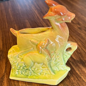 May include: A ceramic deer planter in shades of yellow, orange, and green. The planter features a deer with a fawn, and a rectangular opening for plants. The deer has a glossy finish and is set against a backdrop of leaves.