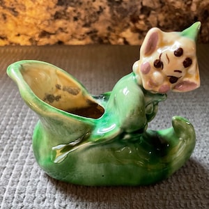 Puede incluir: Un macetero de cerámica verde con forma de zapato con un personaje de dibujos animados caprichoso asomándose por la parte superior. El personaje tiene una cara blanca con ojos negros y mejillas rosadas.