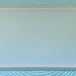May include: A rectangular, light blue plastic container with a matching lid. The container is set on a light blue woven placemat. The lid has a slightly rounded edge and a small circular design in the center.