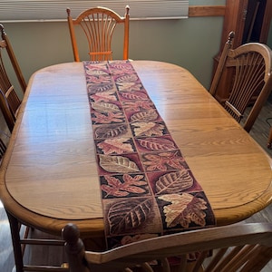 May include: A rectangular table runner featuring a repeating grid pattern of autumn leaves in shades of burgundy, brown, gold, and tan. The runner is centered on a light brown wooden dining table surrounded by several wooden chairs with spindle backs.