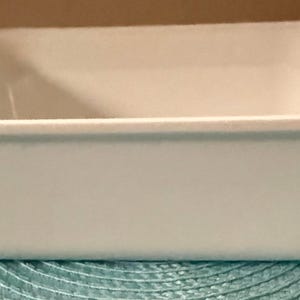May include: A rectangular, light blue baking dish with rounded corners and a slightly flared rim. The dish is made of a solid, opaque material and appears to be empty. The dish is sitting on a teal-colored placemat.