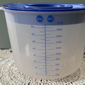 May include: A translucent plastic measuring pitcher with a blue lid and handle. The exterior features blue printed measurement markings in ounces ranging from 8 to 64 and milliliters from 200 to 2000, with a pouring spout on the right side of the rim.