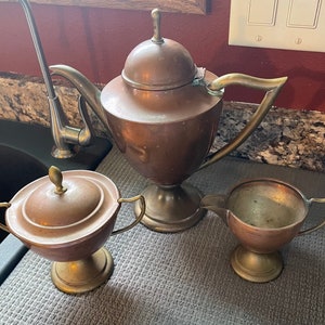 May include: A copper tea set with a teapot, creamer, and sugar bowl. The teapot has a rounded body with a lid and a spout. The creamer and sugar bowl have a round body with a lid and a handle.