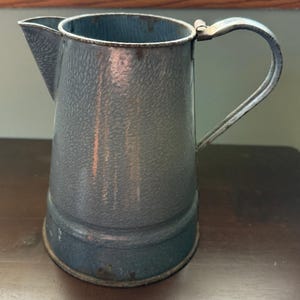 May include: A vintage blue enamel pitcher with a textured surface and a curved handle. The pitcher has a spout for pouring and a metal handle attached to the side. The pitcher is likely used for holding liquids.